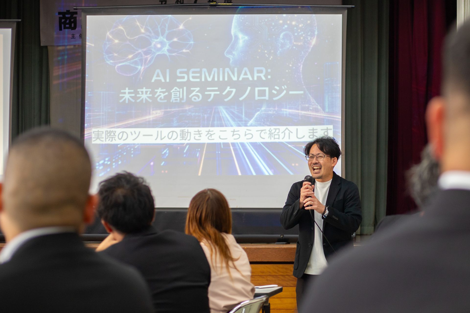 U-WAN代表 棚原秀樹。AIセミナーで登壇し、マイクを手に参加者へ笑顔で語りかけている様子。